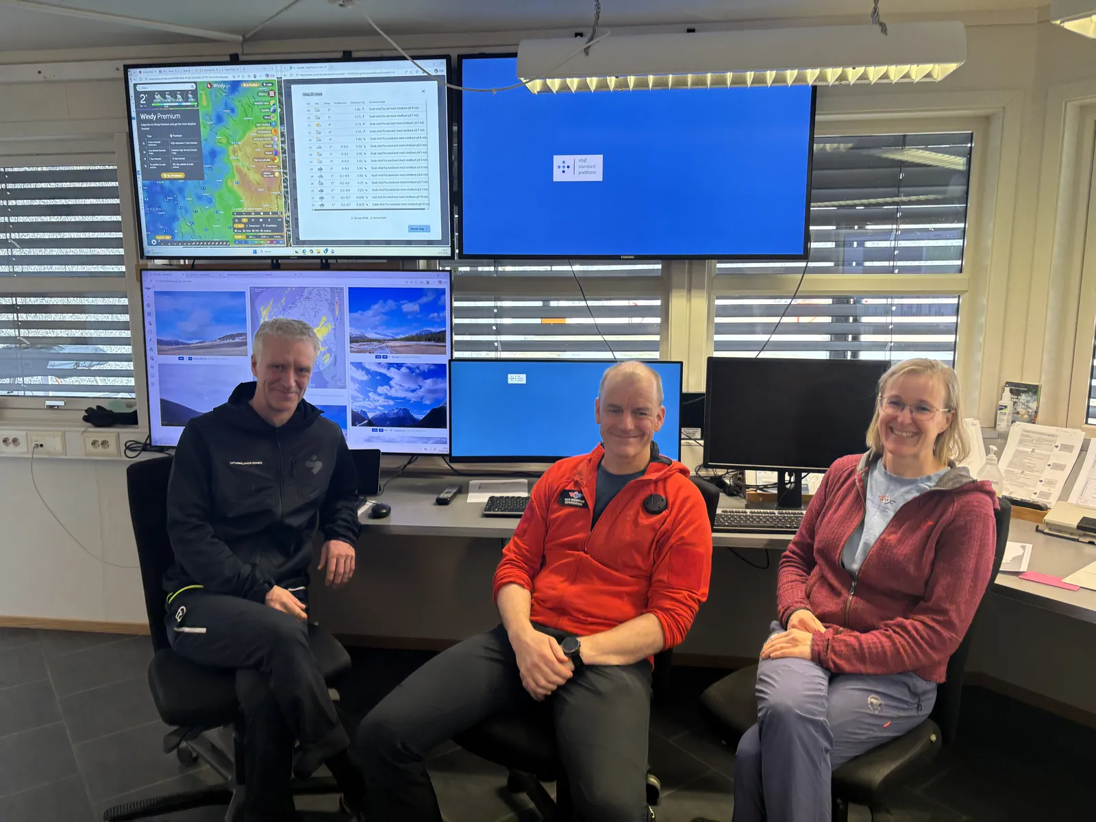 To menn og en dame foran mange skjermer