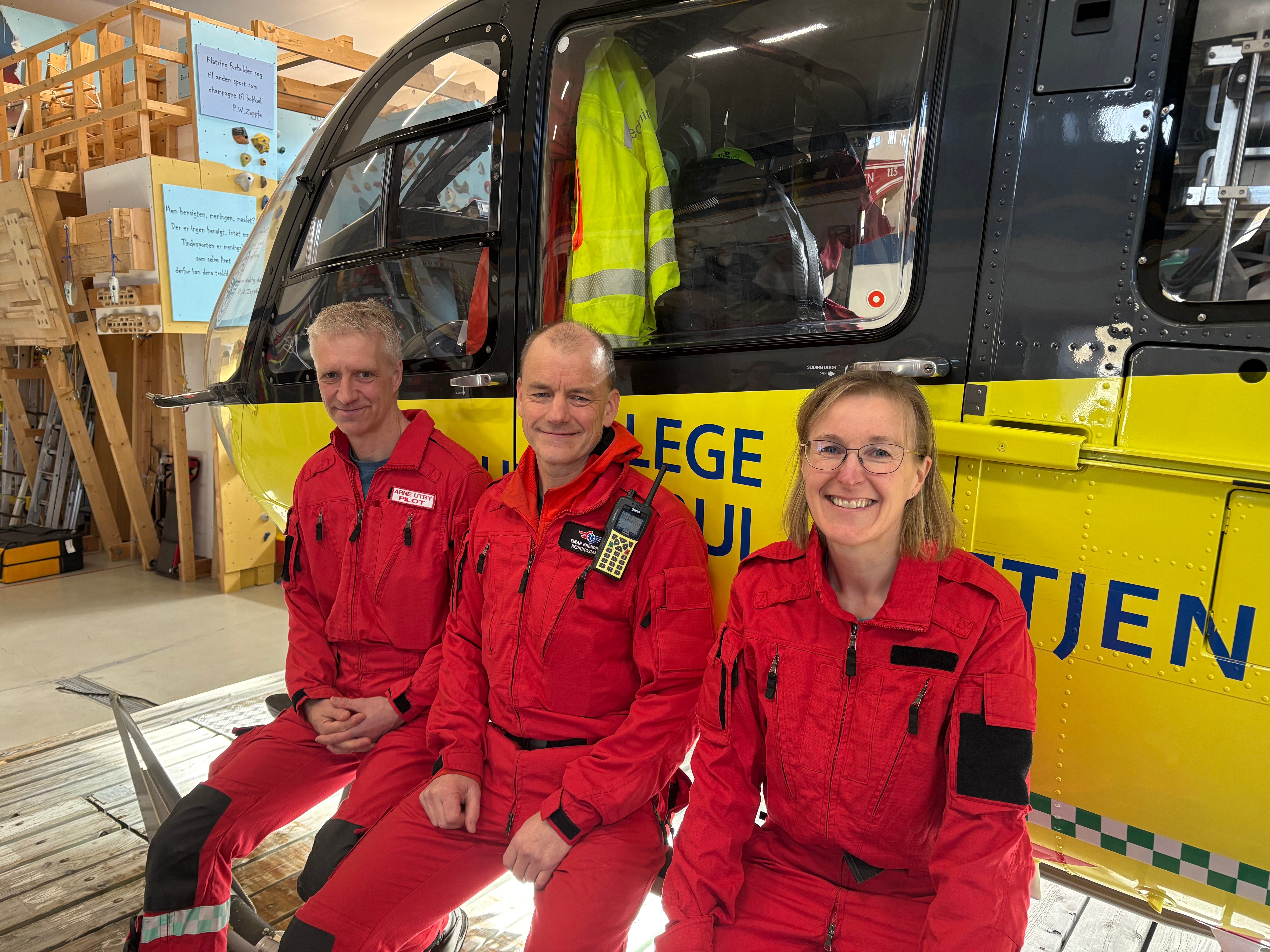 To menn og en dame i røde  luftambulanseuniformer foran et gult helikopter