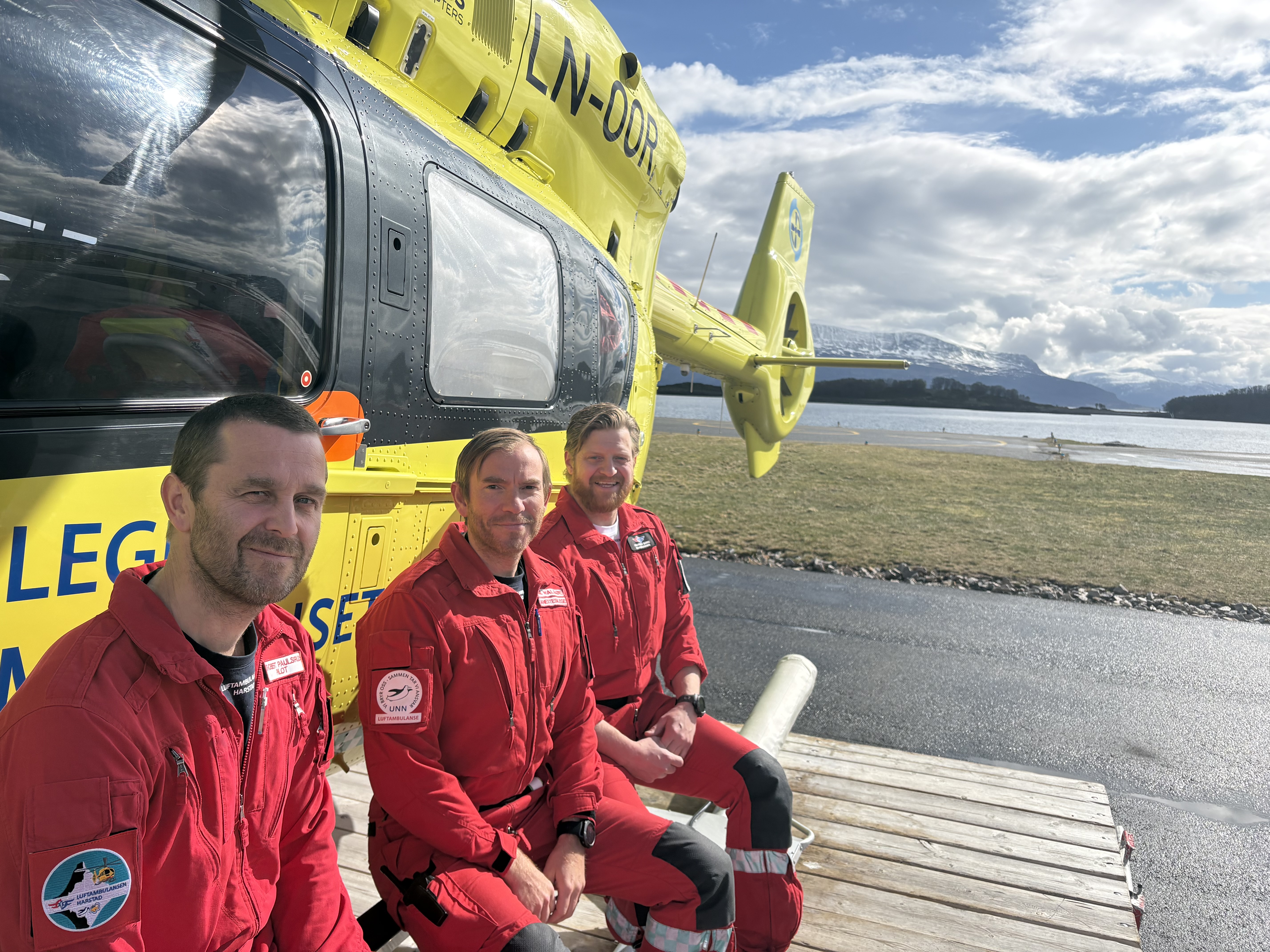 Tre menn i røde uniformer foran et gult helikopter
