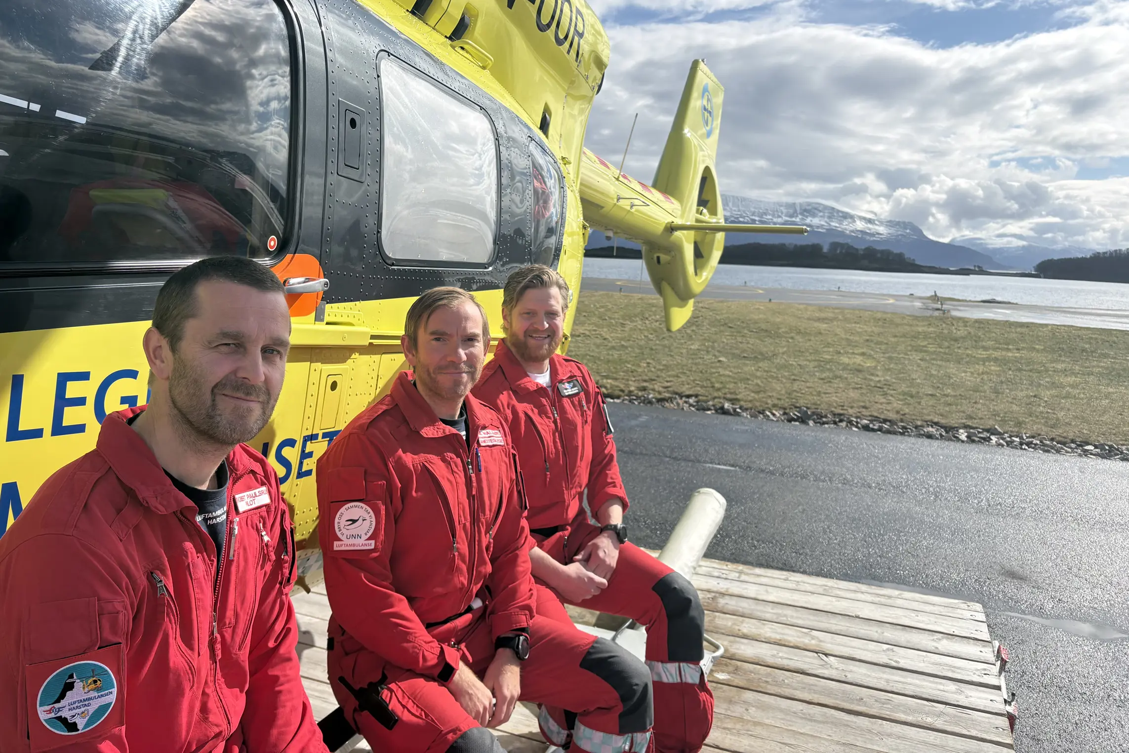 Tre menn i røde uniformer foran et gult helikopter