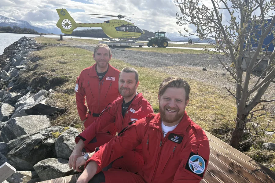Tre smilende menn i røde uniformer med et gult helikopter i bakgrunnen