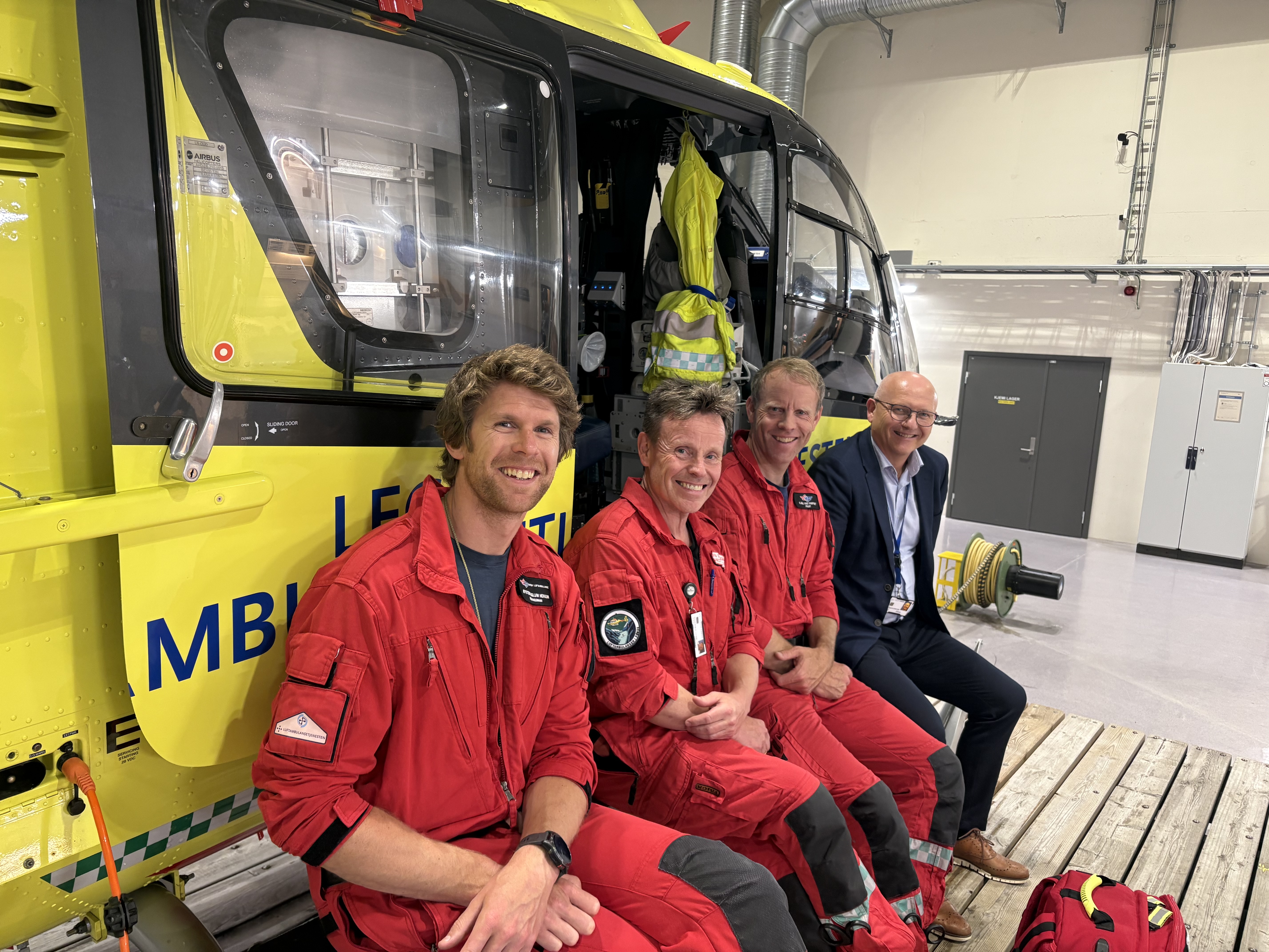 Tre menn i røde uniformer og en mann i dress foran et gult ambulansehelikopter