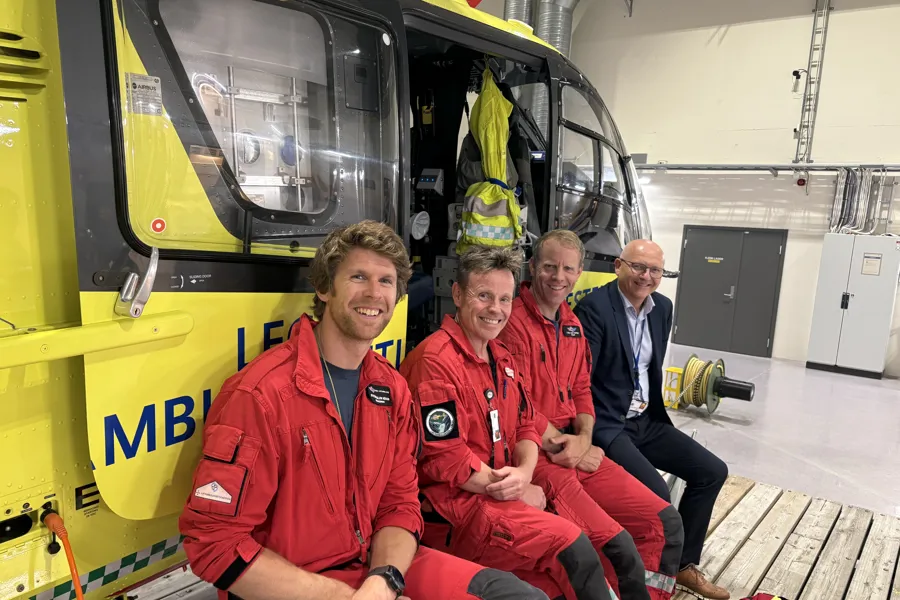 Tre menn i røde uniformer og en mann i dress foran et gult ambulansehelikopter