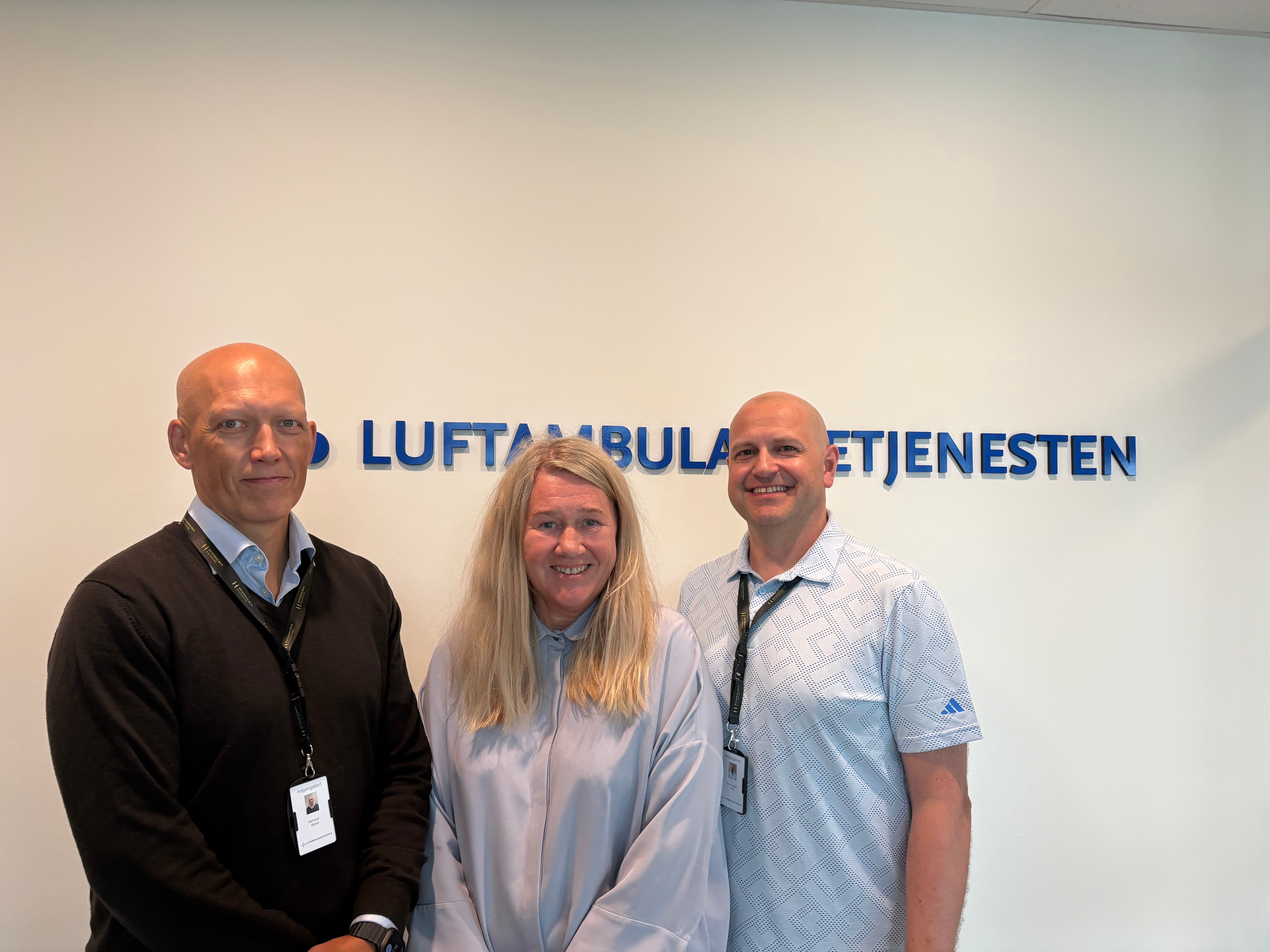 To menn og e kvinne foran Luftambulansetjenestens logo
