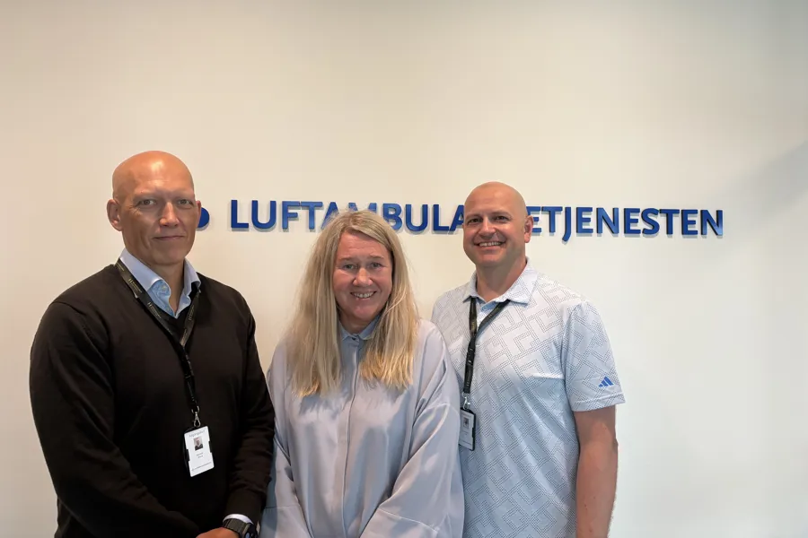 To menn og e kvinne foran Luftambulansetjenestens logo