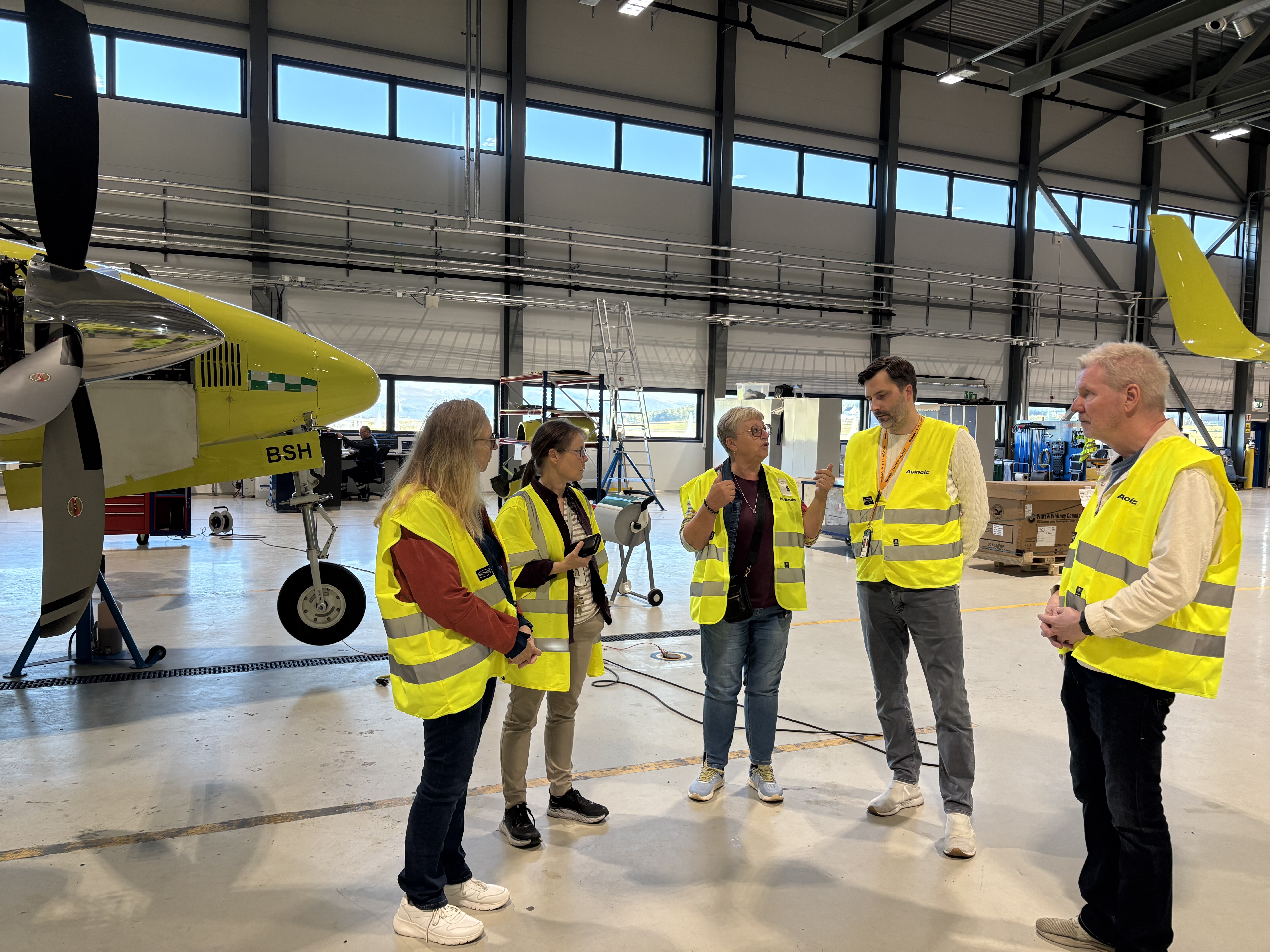 Tre damer og tre menn i refleksvest inne i en flyhangar